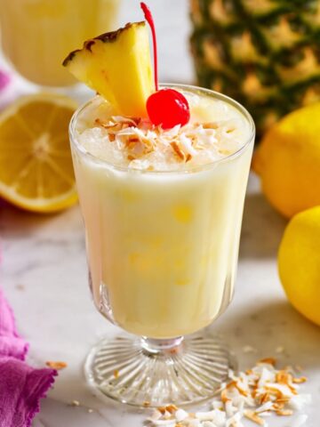 A creamy pale yellow coconut pineapple lemonade in a footed glass, topped with toasted coconut flakes, a maraschino cherry, and a fresh pineapple wedge, surrounded by whole lemons, a halved lemon, and a fresh pineapple on a marble surface.