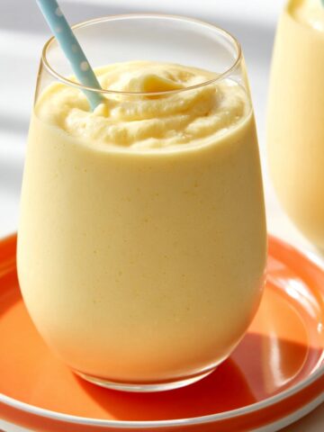 A thick creamy pale yellow creamsicle slushy in a stemless glass with a light blue polka dot straw, served on an orange plate, with a second glass visible in the background.