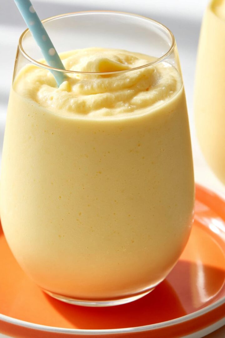 A thick creamy pale yellow creamsicle slushy in a stemless glass with a light blue polka dot straw, served on an orange plate, with a second glass visible in the background.