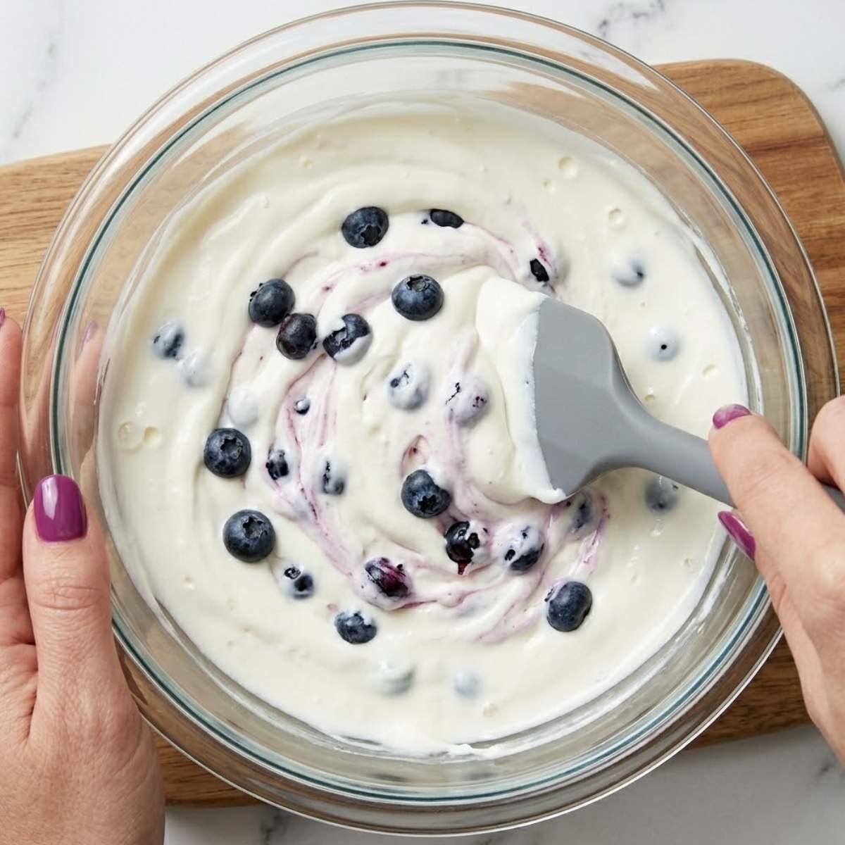 Extreme close-up view of hands with purple nails using a silicone spatula to gently mix whole fresh blueberries into smooth white cream cheese.