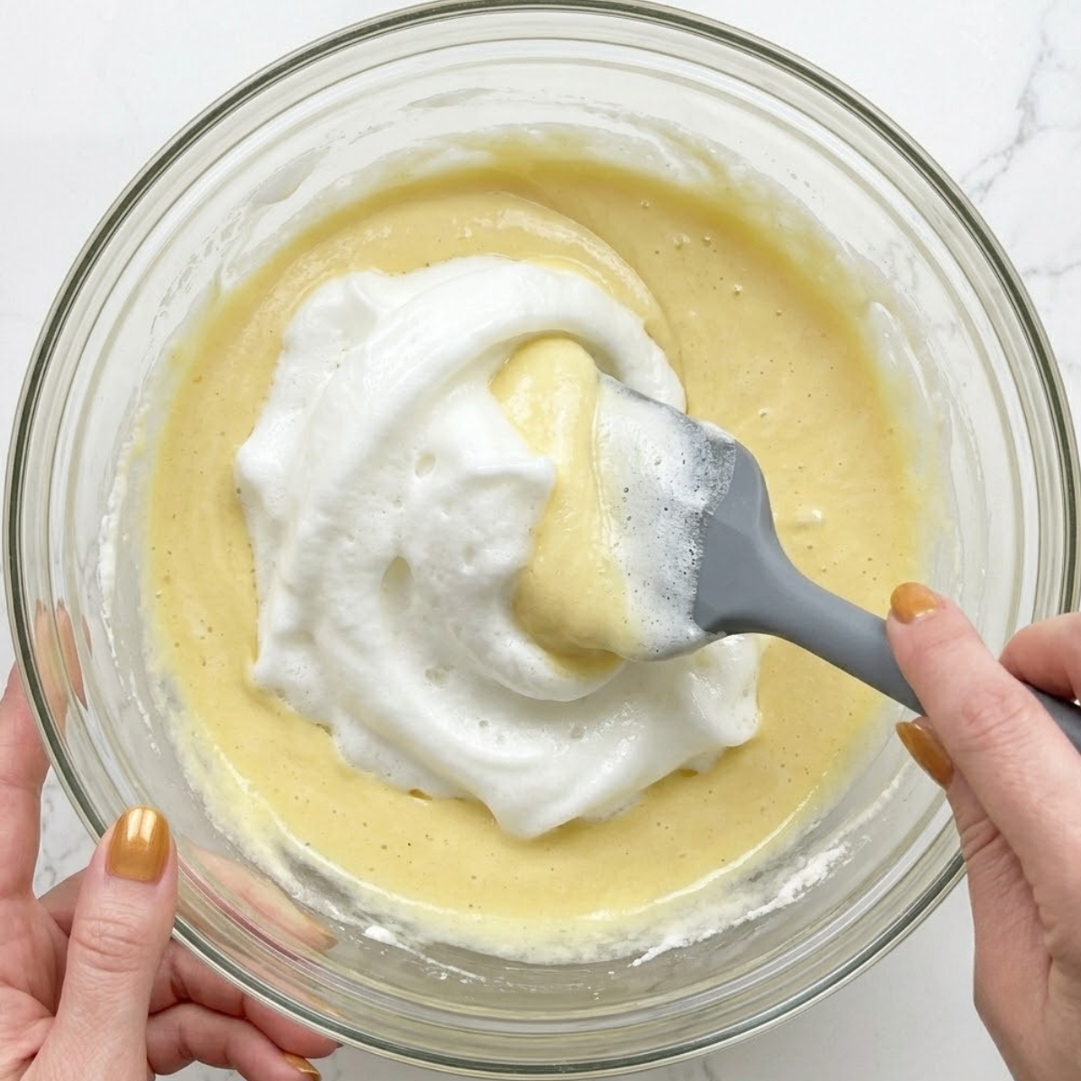 Extreme close-up view of hands with gold nails using a silicone spatula to gently fold whipped egg whites into a soufflé pancake batter base.