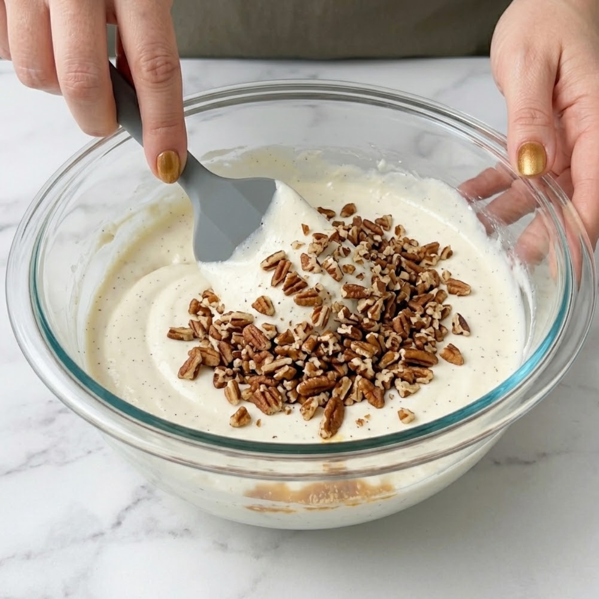 Tight close-up zoom of hands with amber nails using a silicone spatula to fold toasted chopped pecans into thick white cheesecake batter.