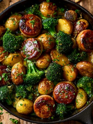 A cast iron skillet filled with caramelized kielbasa rounds, roasted baby potatoes, and tender broccoli florets in a glossy honey garlic sauce, garnished with fresh chopped herbs on a rustic wooden surface.