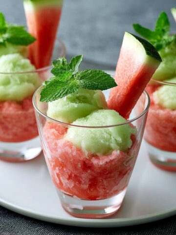 Three glass cups layered with pink salted watermelon granita and pale green ice cream scoops, garnished with fresh mint sprigs and a watermelon wedge, arranged on a white plate.