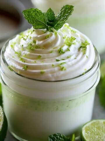 A glass jar filled with pale green yogurt lime mousse, topped with a swirl of whipped cream, fresh lime zest, and a sprig of mint, surrounded by halved limes and mint leaves.