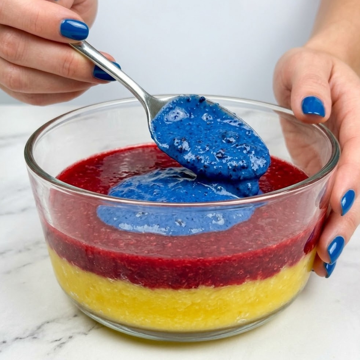 Tight close-up zoom of hands with blue nails spooning a vibrant sapphire blue layer onto a multi-layered tropical fruit colada.