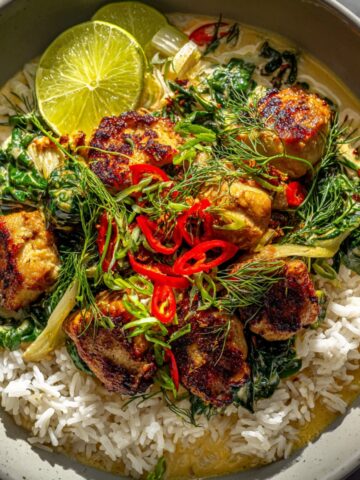 A bowl of lemongrass chicken in creamy Thai coconut curry with wilted greens, served over steamed white rice, garnished with sliced red chilies, fresh dill, green onions, and a lime wedge.