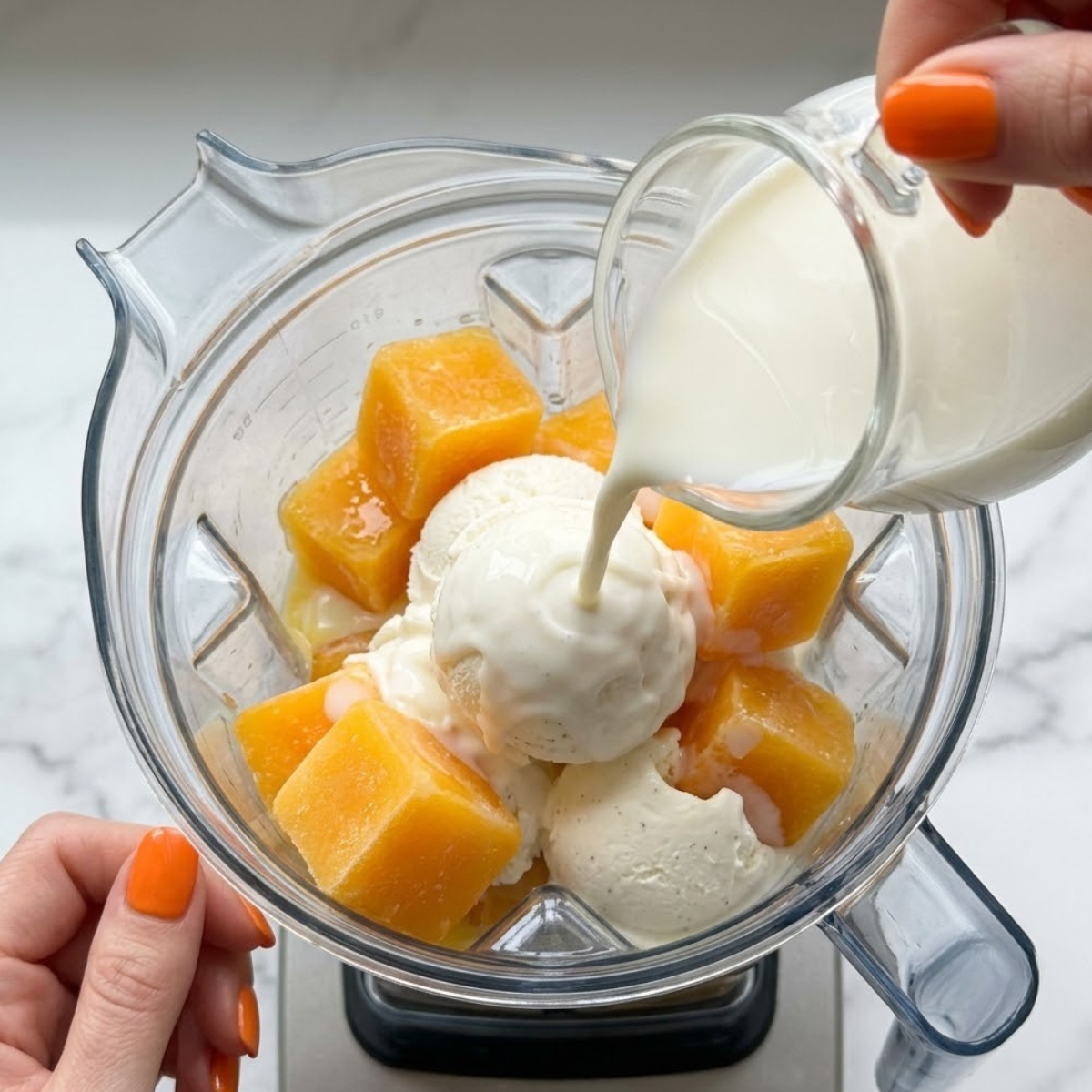 Extreme close-up view of hands with orange nail polish pouring milk over frozen orange juice cubes and vanilla ice cream inside a blender.