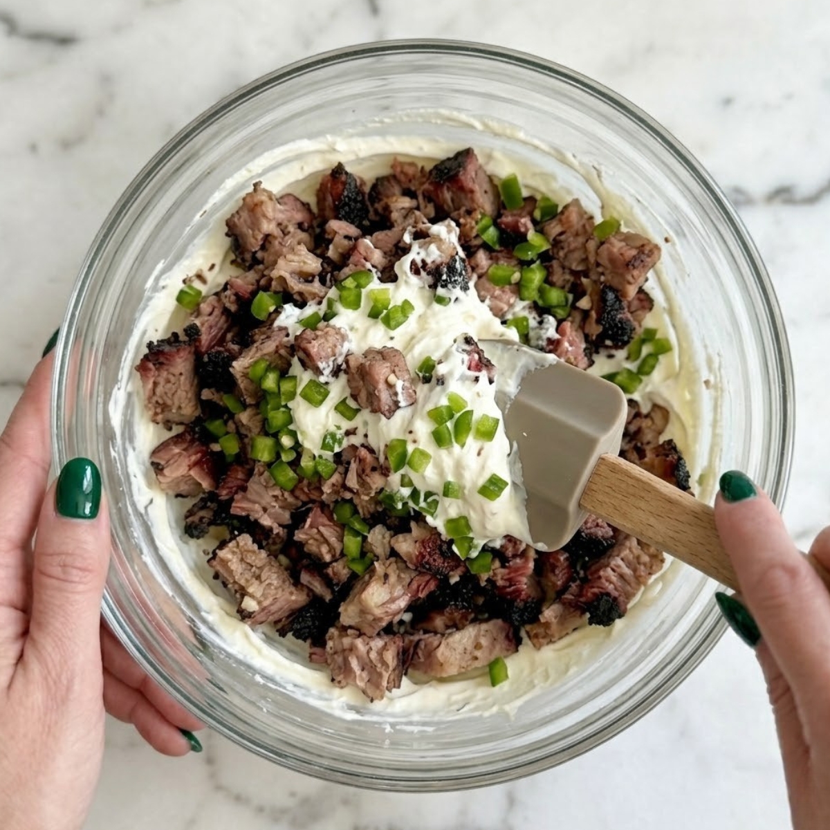 Extreme close-up of hands with emerald green nails mixing chopped brisket and diced jalapeños into cream cheese with a silicone spatula.
