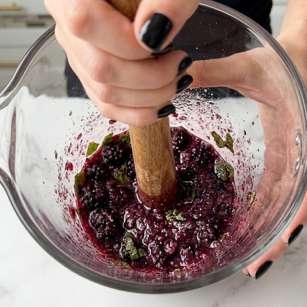 Extreme close-up view of hands with glossy black nails using a wooden muddler to mash blackberries, sugar, and mint in a glass pitcher.