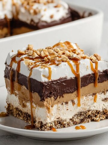 A layered slice of peanut butter chocolate pretzel pie with a crunchy pretzel crust, cream cheese layer, peanut butter mousse, rich chocolate ganache, and whipped cream, drizzled with caramel sauce and topped with chopped peanuts, served on a white plate.