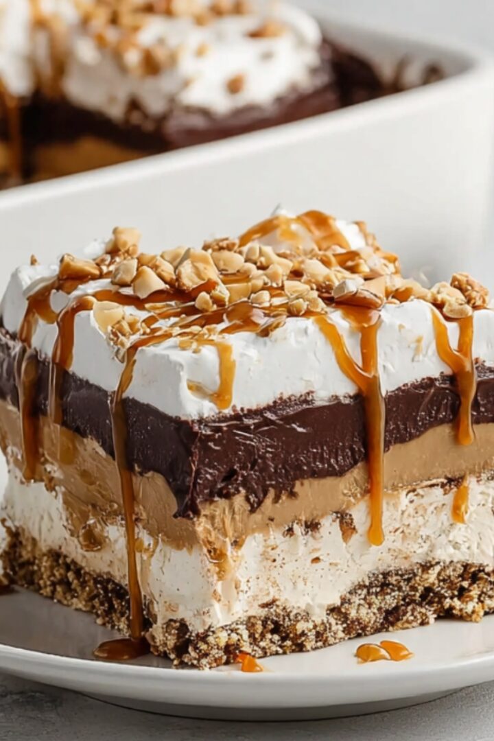A layered slice of peanut butter chocolate pretzel pie with a crunchy pretzel crust, cream cheese layer, peanut butter mousse, rich chocolate ganache, and whipped cream, drizzled with caramel sauce and topped with chopped peanuts, served on a white plate.