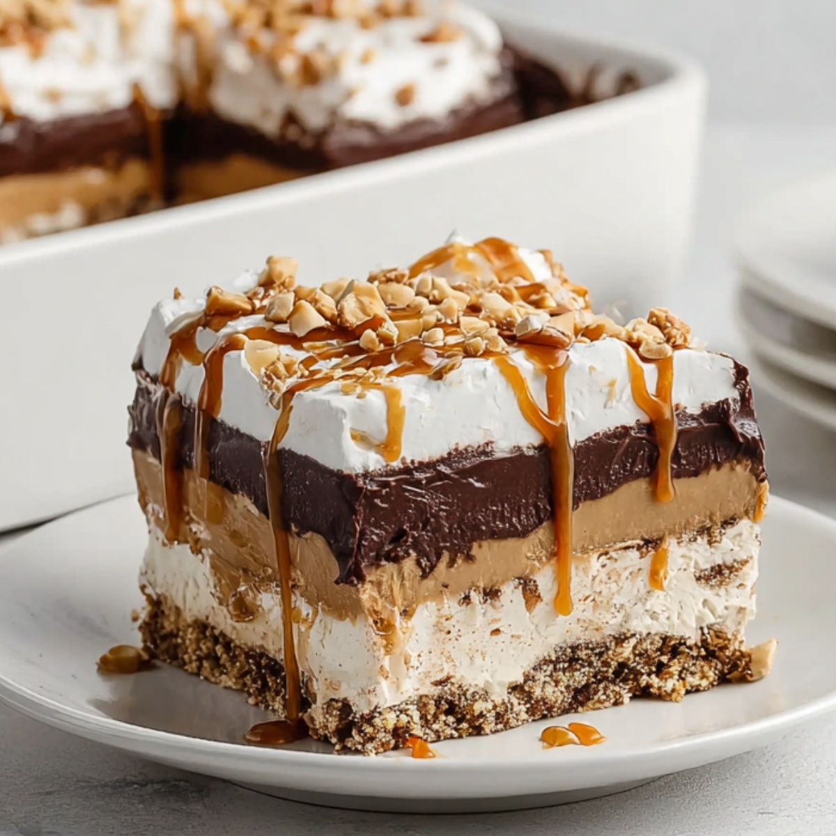 A layered slice of peanut butter chocolate pretzel pie with a crunchy pretzel crust, cream cheese layer, peanut butter mousse, rich chocolate ganache, and whipped cream, drizzled with caramel sauce and topped with chopped peanuts, served on a white plate.