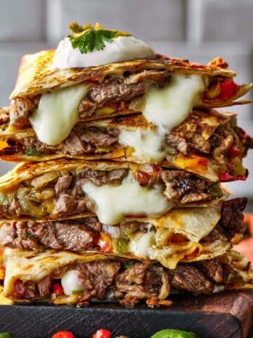 A stack of crispy Philly cheesesteak quesadillas filled with sliced steak, sautéed peppers and onions, and melted provolone, topped with a dollop of sour cream and fresh cilantro.