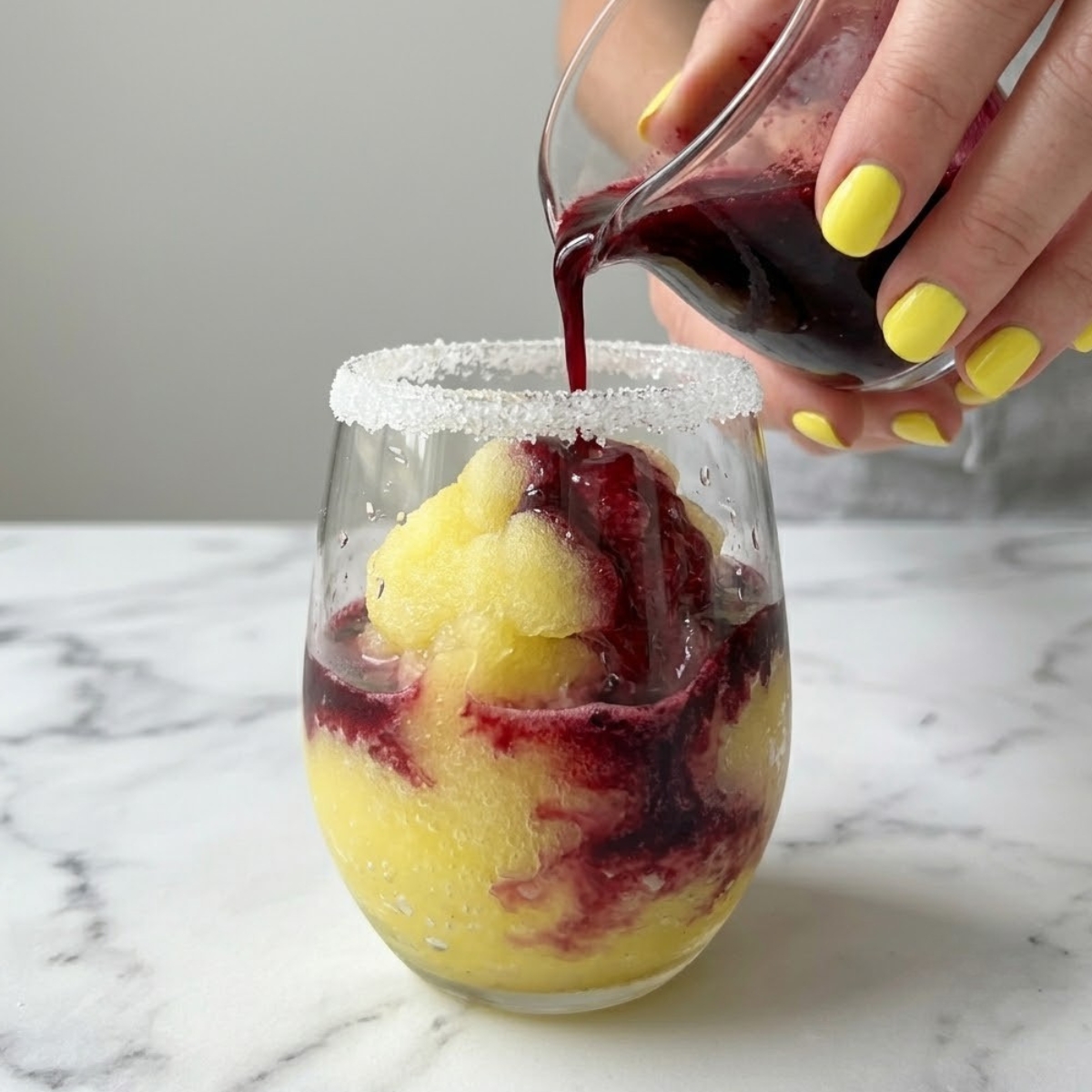 Tight close-up zoom of hands with yellow nails slowly layering a dark purple blueberry reduction over a thick yellow lemon slush inside a sugar-rimmed glass.