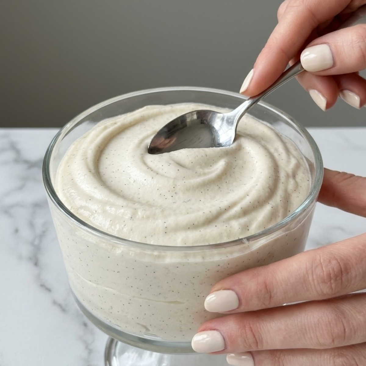 Tight close-up zoom of hands with cream-colored nails using a spoon to create a decorative swirl on top of a vanilla mousse cup.