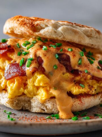 A toasted English muffin breakfast sandwich piled high with fluffy scrambled eggs and crispy bacon, smothered in creamy chipotle sauce, garnished with chopped chives and smoked paprika, served on a speckled ceramic plate.