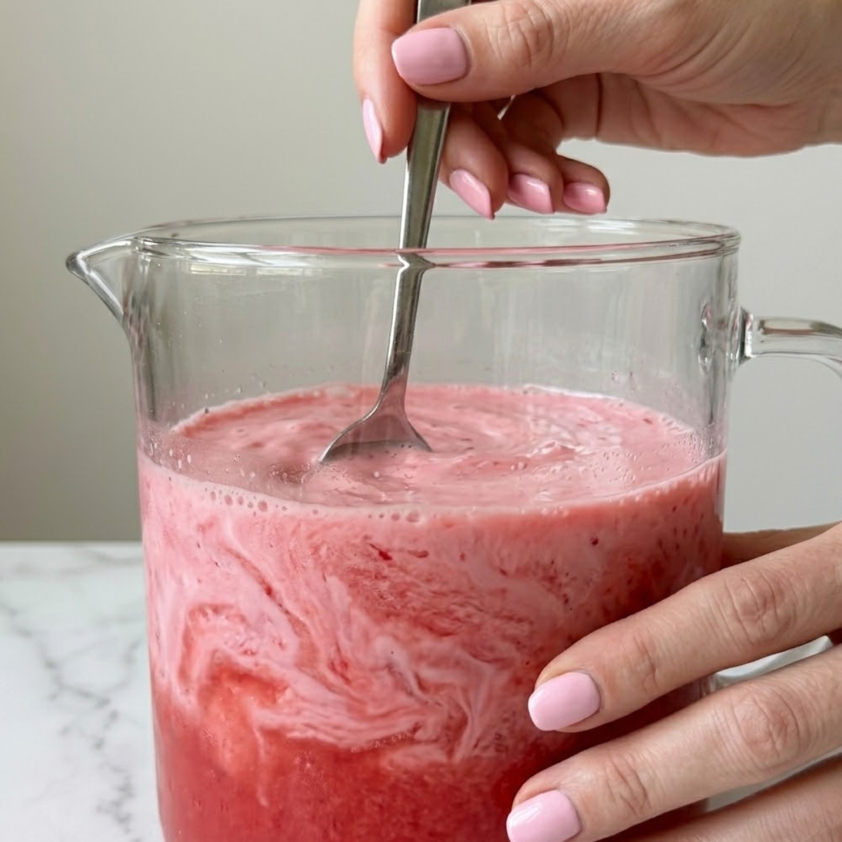 Tight close-up zoom of hands with pink nails using a long silver spoon to mix fresh watermelon rose drink inside a glass pitcher.