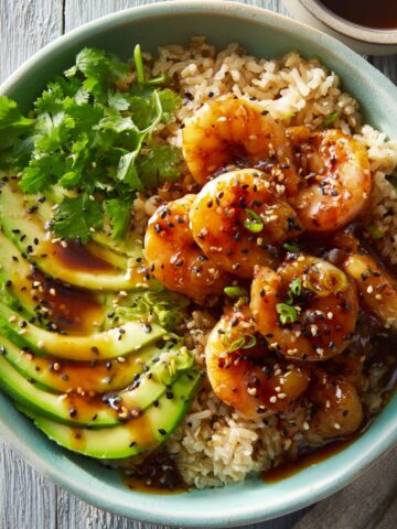 A teriyaki shrimp rice bowl in a teal ceramic bowl with glazed shrimp, sliced avocado, and brown rice, topped with black and white sesame seeds, sliced green onions, and fresh cilantro, with teriyaki sauce drizzled throughout.