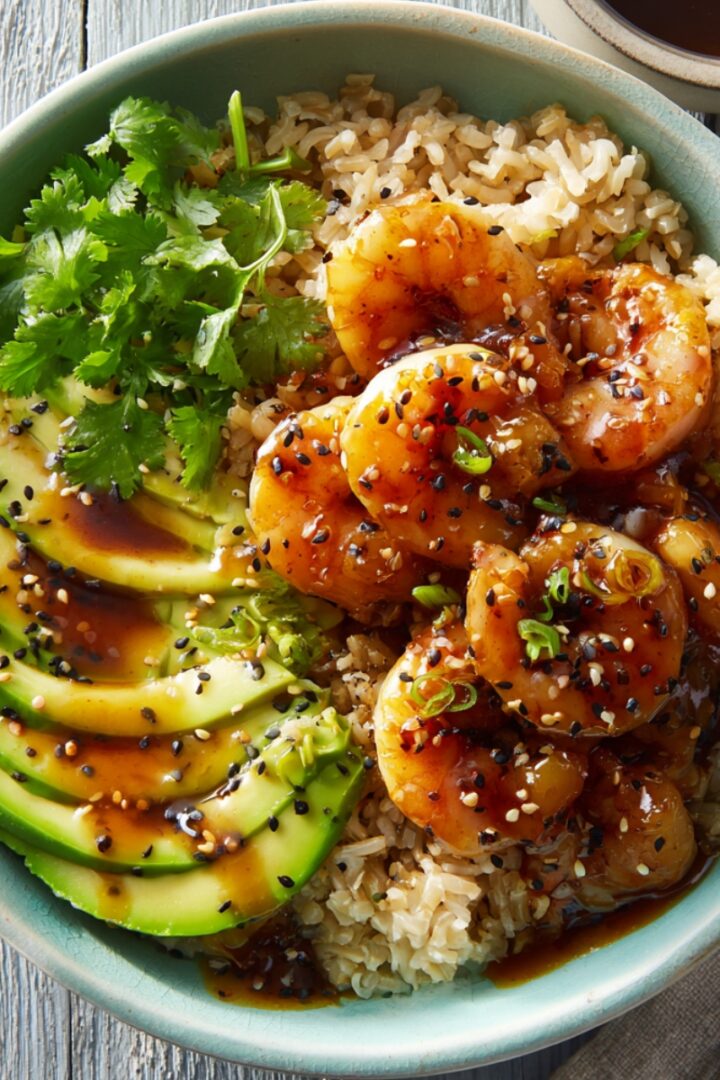 A teriyaki shrimp rice bowl in a teal ceramic bowl with glazed shrimp, sliced avocado, and brown rice, topped with black and white sesame seeds, sliced green onions, and fresh cilantro, with teriyaki sauce drizzled throughout.