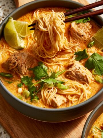 A bowl of Thai red curry noodle soup with chunks of chicken, egg noodles being lifted with chopsticks, fresh cilantro, green onions, and lime wedges in a creamy orange coconut curry broth.