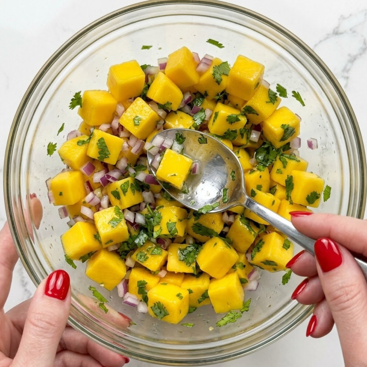 Extreme close-up view of hands with red nails using a spoon to gently toss cubed mango, red onion, and cilantro inside a glass bowl.
