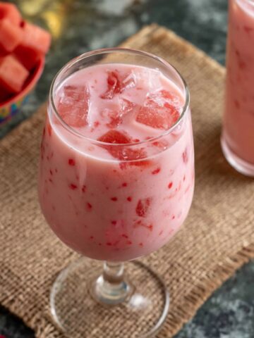 A pink, creamy watermelon rose drink served in a stemmed glass with chunks of fresh watermelon, set on a burlap mat next to a bowl of cubed watermelon.