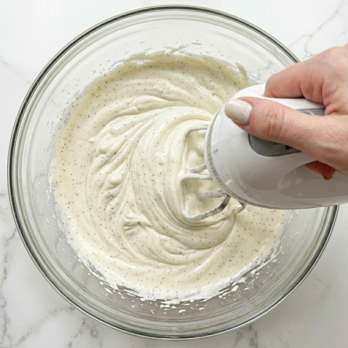 Extreme close-up view of hands with cream-colored nails using a hand mixer to whip heavy cream with vanilla bean paste inside a glass bowl.
