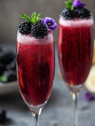Two elegant champagne flutes filled with a deep red sparkling blackberry mojito mocktail, garnished with fresh blackberries, mint sprigs, and purple edible flowers, with a bowl of blackberries and lemon halves in the background.