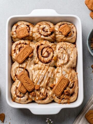 A high-angle close-up of Biscoff cinnamon roll inside a ceramic dish with crushed biscuit garnish on a marble counter.