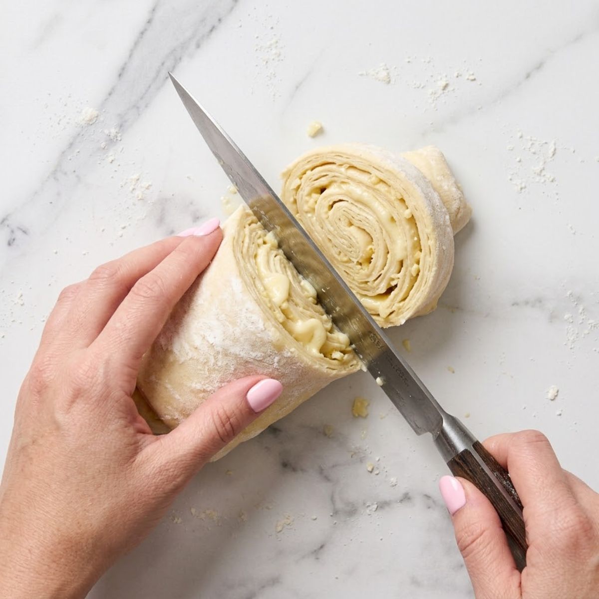 Extreme close-up shot from above showing a woman with baby pink nails using a chef's knife to slice through a rolled puff pastry dough log, revealing layers of cheese and garlic, on a lightly floured marble counter.