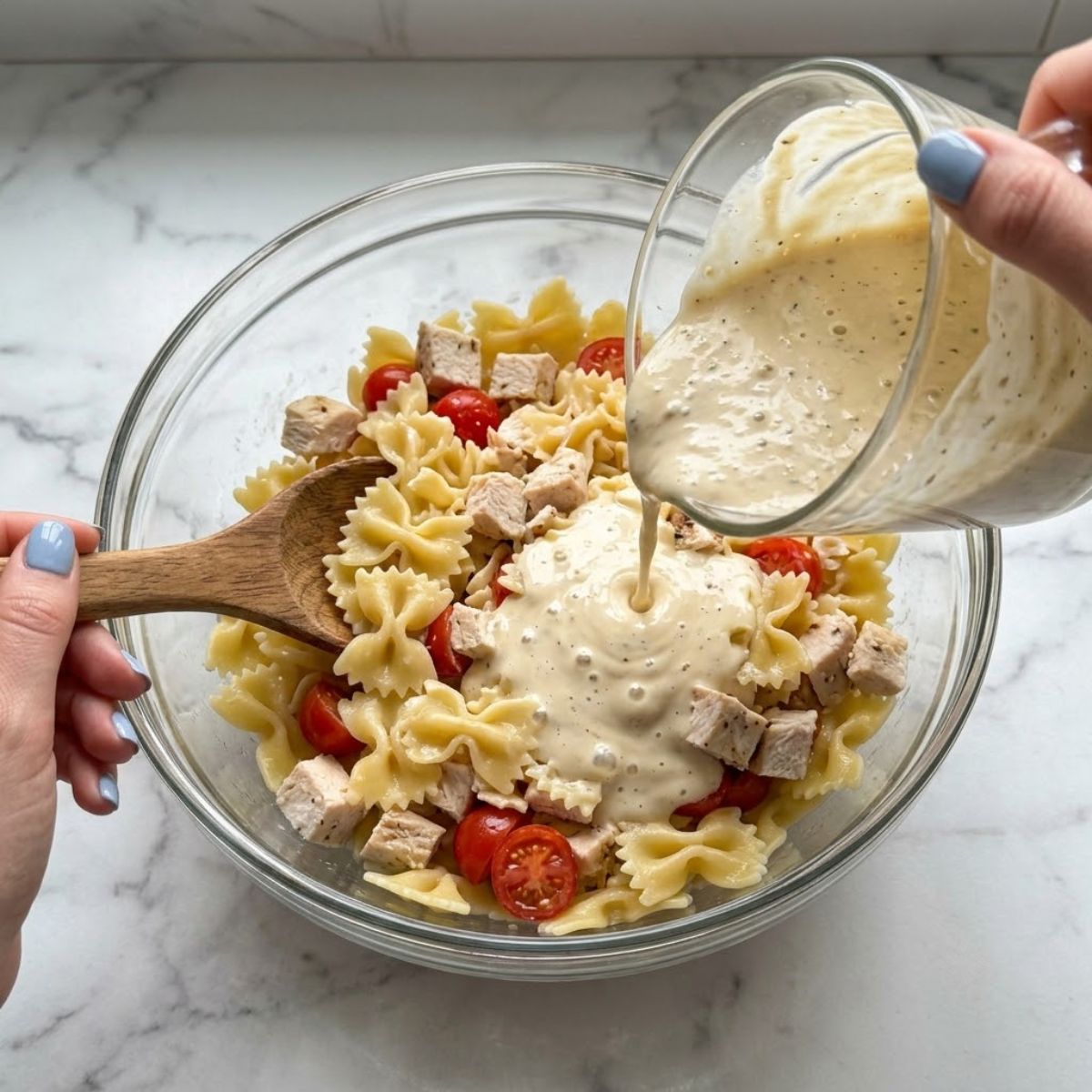 Close-up of hands with blue nails pouring Caesar dressing over pasta and chicken.