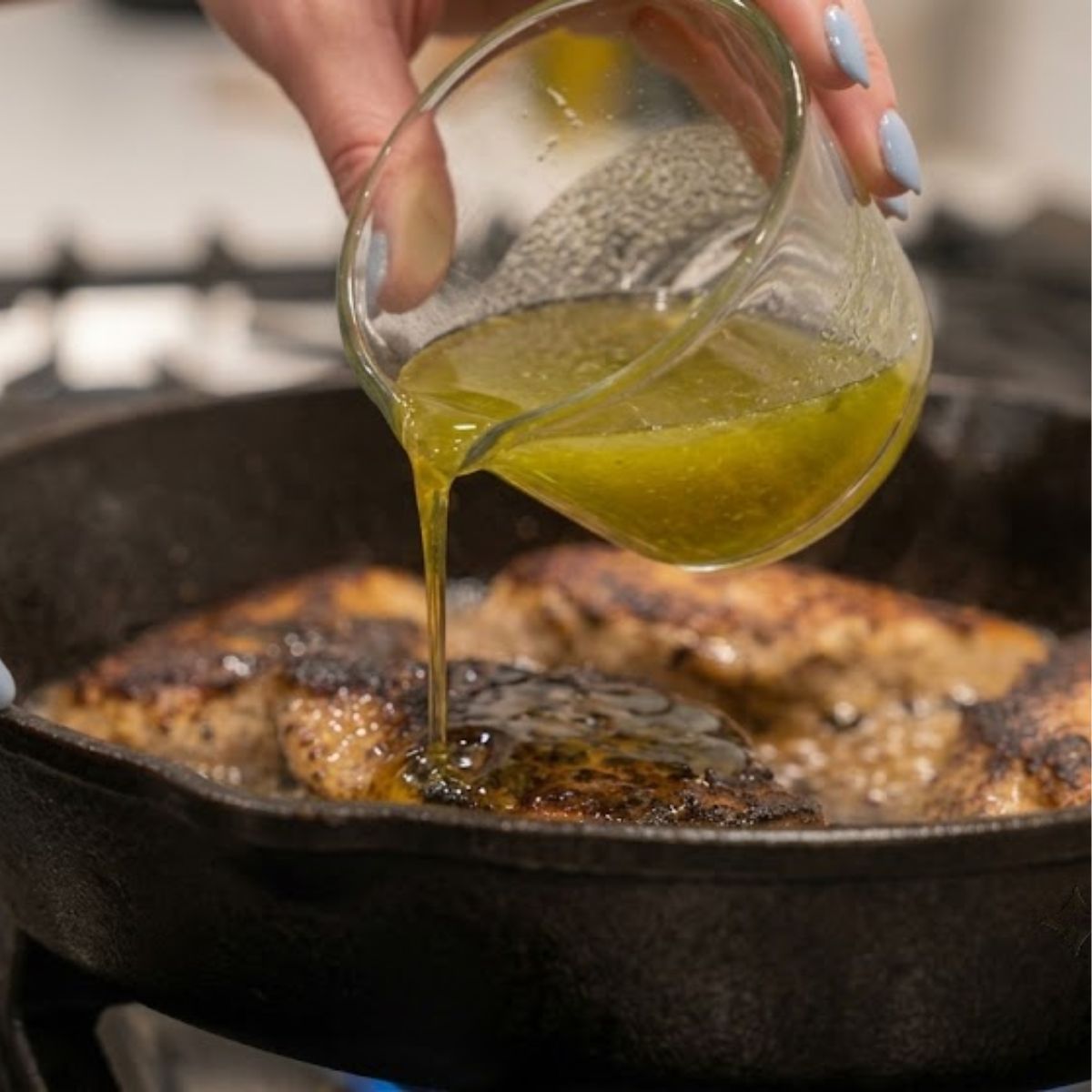 Close-up of hands with blue nails pouring honey and lime juice over chicken in a simmering skillet.