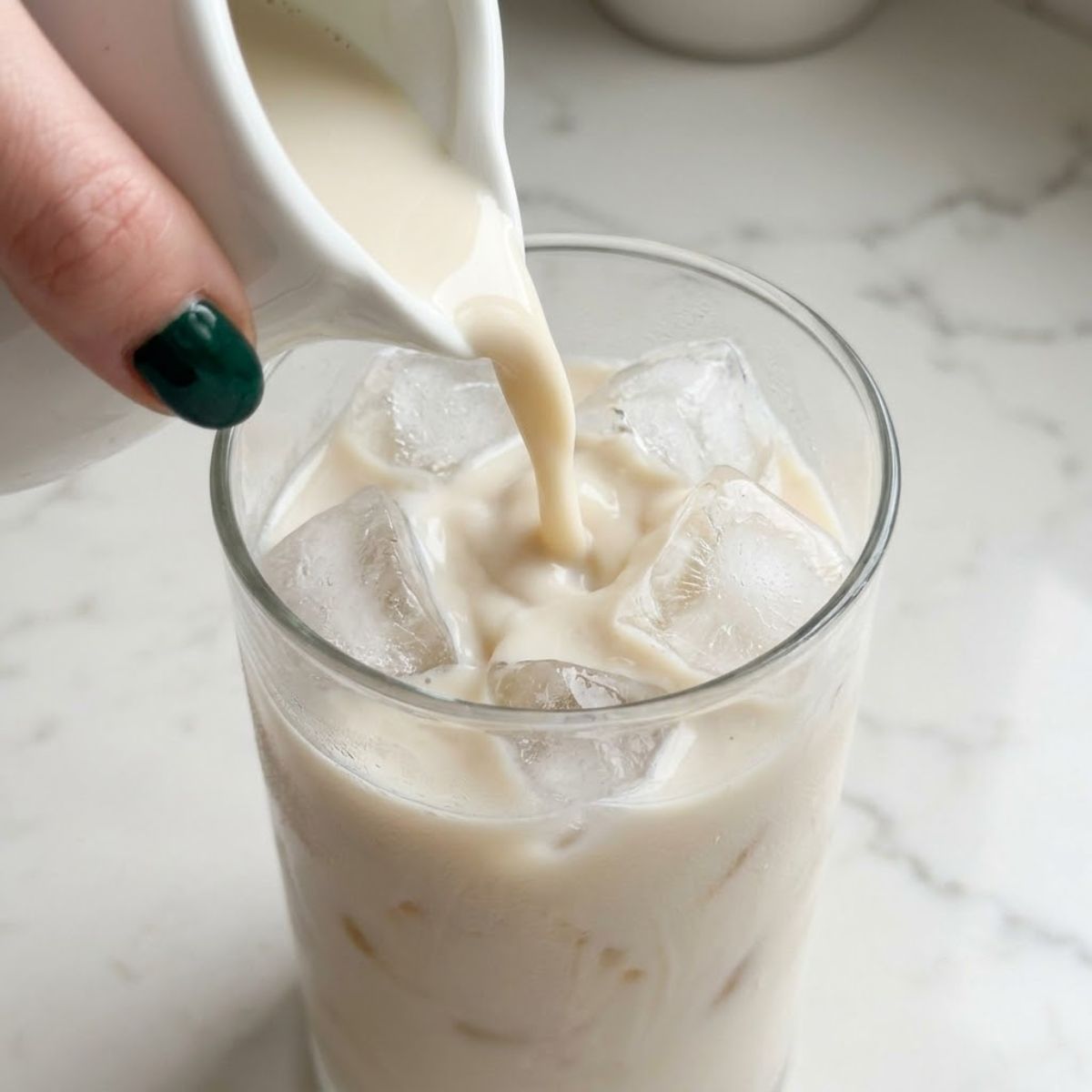 Extreme close-up of a woman with emerald green nails pouring cold oat milk over a tall glass filled with ice cubes on a white marble kitchen counter.