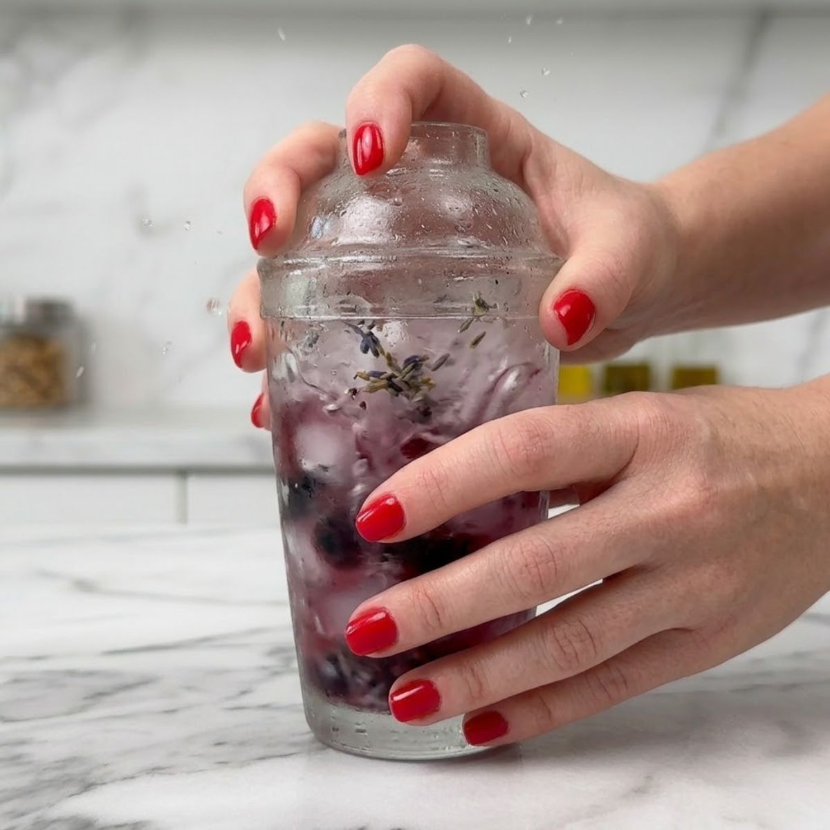 Close-up of hands with red nails vigorously shaking a frost-covered glass cocktail shaker with purple liquid and ice.