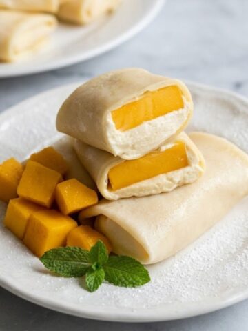 A view of Mango Pancakes with a cut pancake revealing a mango and whiped cream filling on a white ceramic plate on white marble kitchen counter.