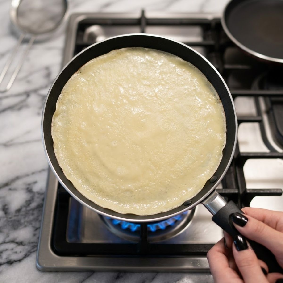 Top down view of cooking a thin yellow crepe in a non-stick pan over a blue gas flame.