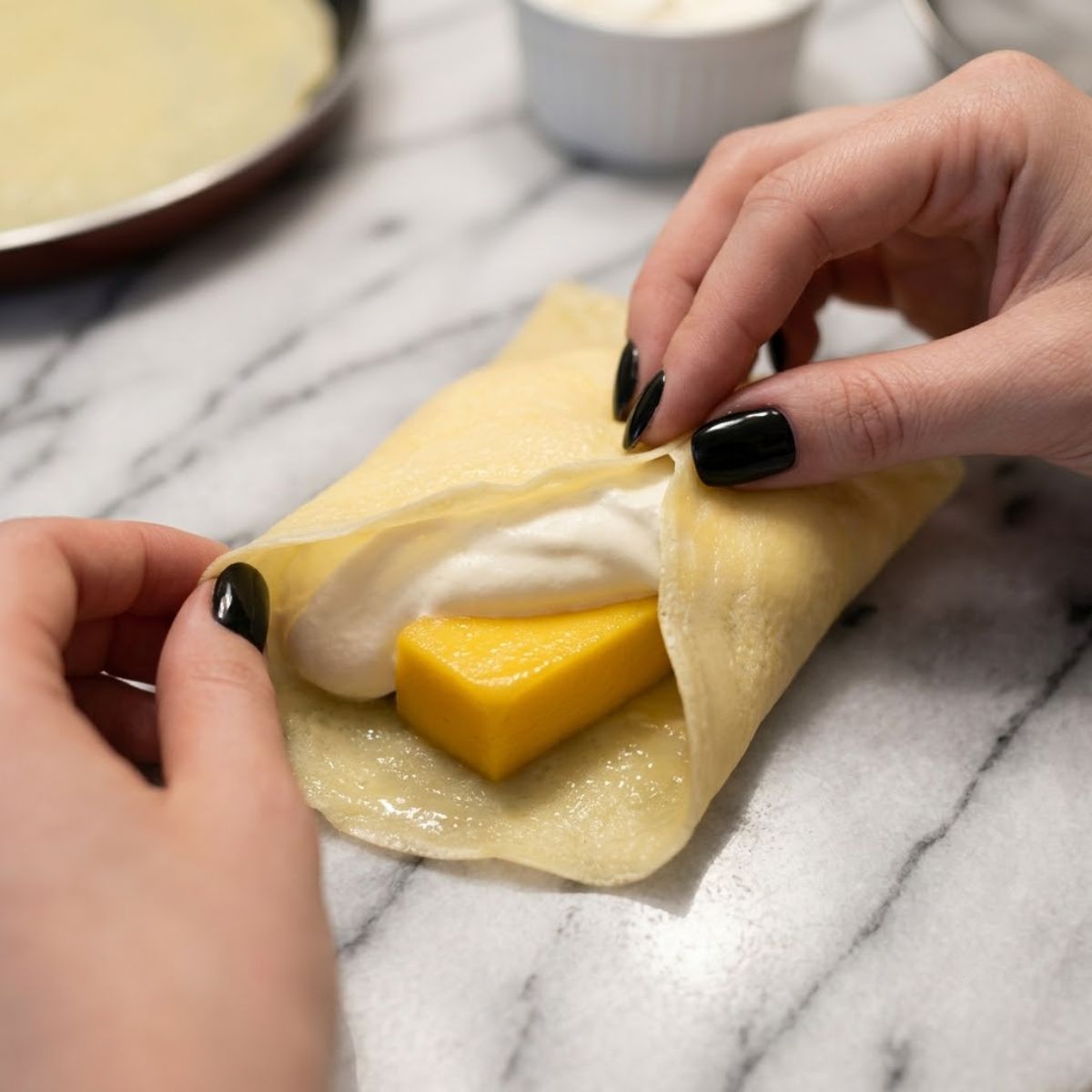 Close-up of hands folding a thin yellow crepe over whipped cream and a sliced mango block on a marble counter.