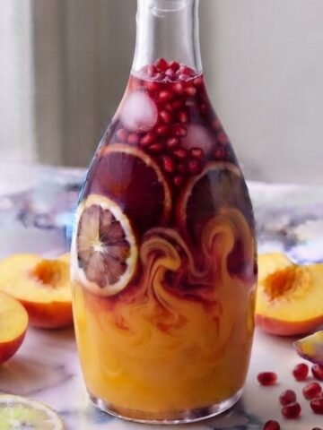 Peach Pomegranate Lemonade contained in a glass bottle on white marble kitchen counter with sliced peaches and pomegranate.