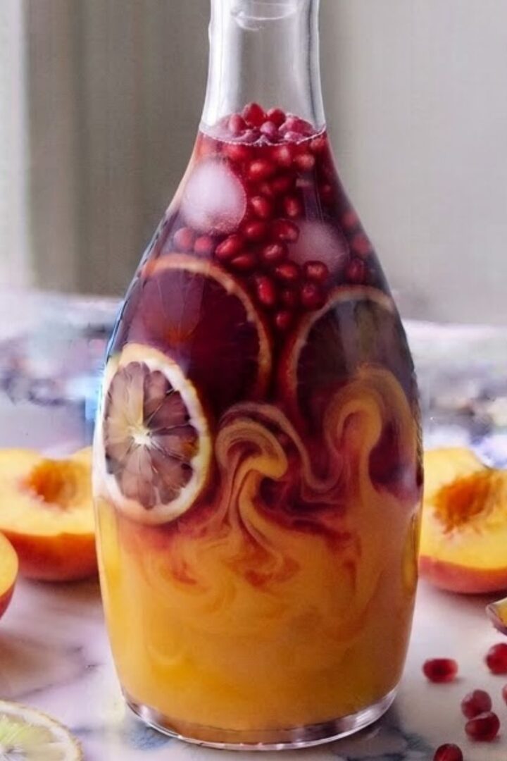 Peach Pomegranate Lemonade contained in a glass bottle on white marble kitchen counter with sliced peaches and pomegranate.