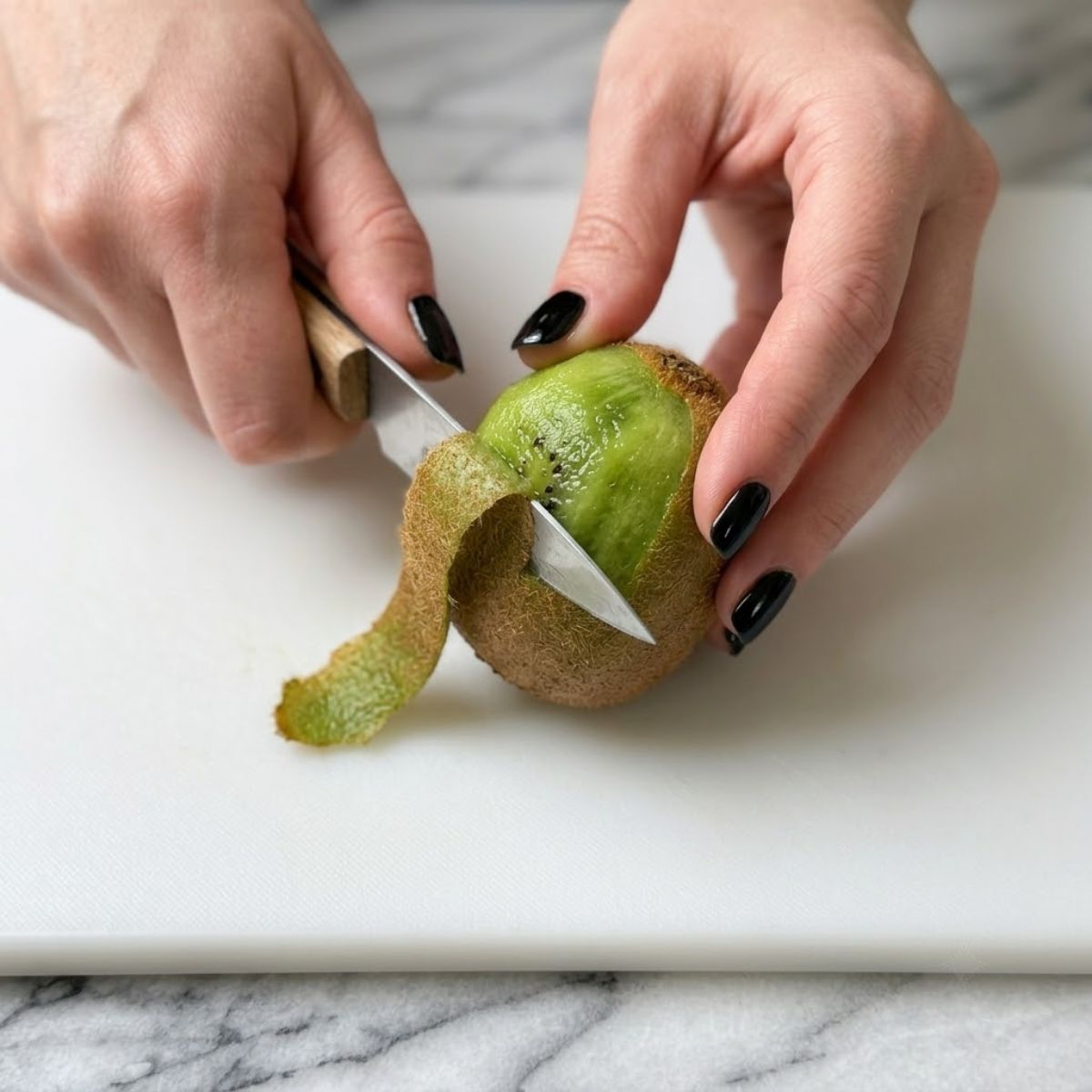 Extreme close-up shot of a woman with glossy black nails peeling a fuzzy fresh kiwi fruit with a paring knife on a cutting board, preparing ingredients for a salad.