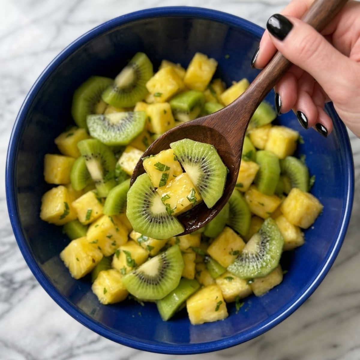 Extreme close-up shot from above showing a woman with glossy black nails gently tossing diced pineapple and sliced kiwi with a wooden spoon inside a blue bowl on a marble counter.