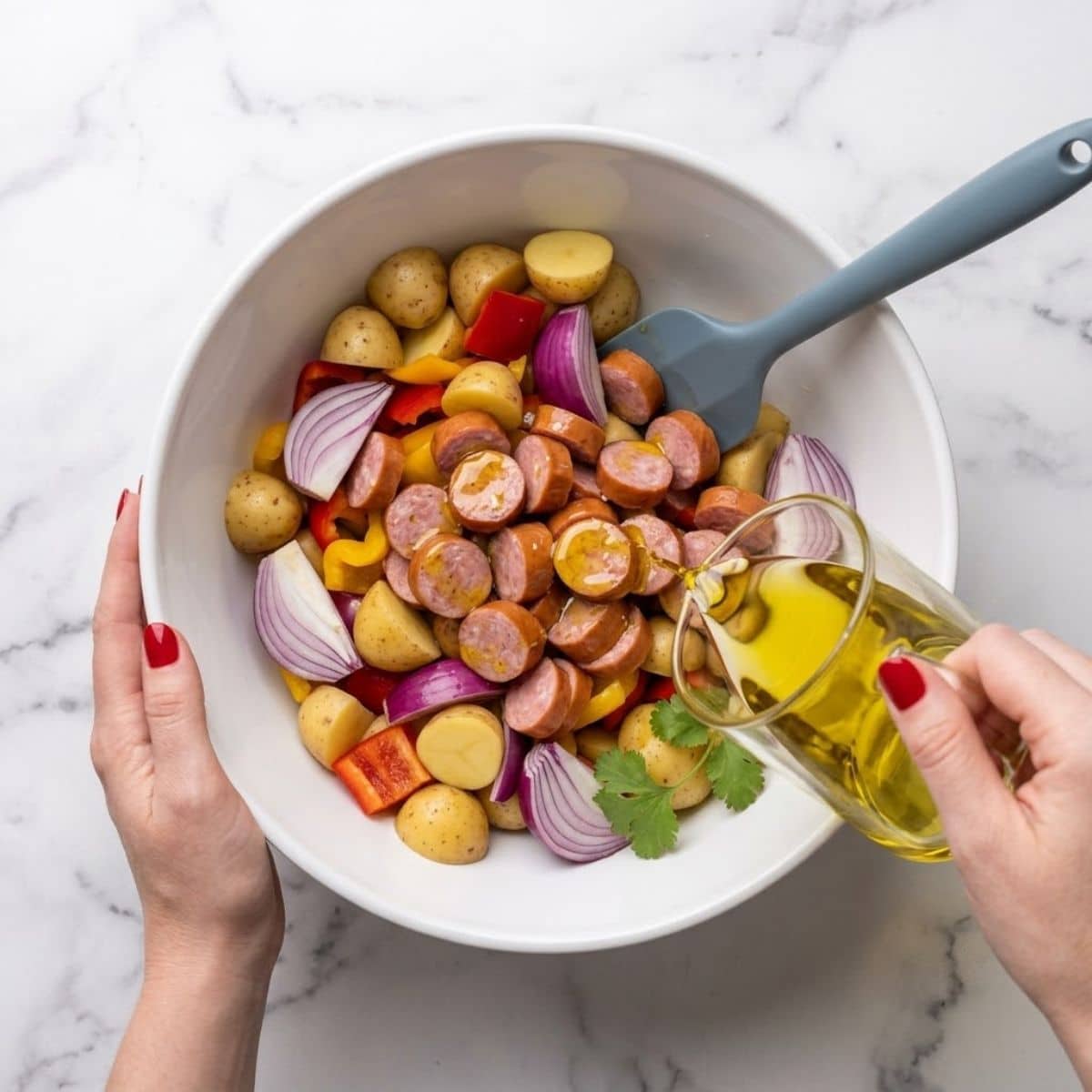 A close-up view of a woman with Classic Red nails drizzling olive oil over raw sausage, potatoes, and vegetables in a mixing bowl on a marble counter.