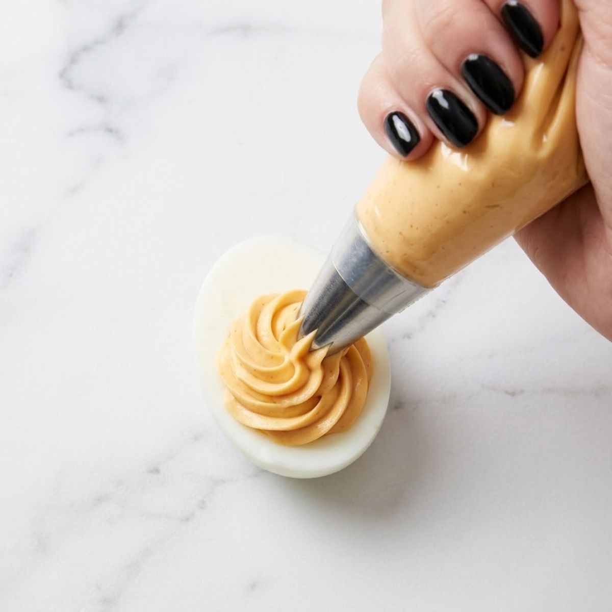 Extreme close-up shot from above showing a woman with glossy black nails piping a smooth yolk filling swirl into a halved boiled egg white on a marble counter.