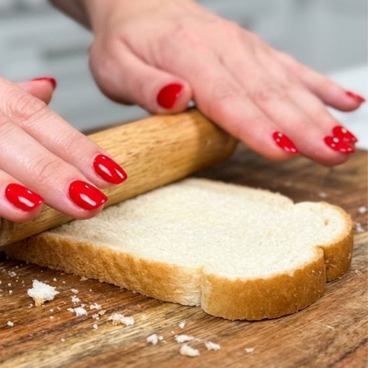 Close-up of hands with red nails using a rolling pin to flatten a slice of white bread on a wooden board.