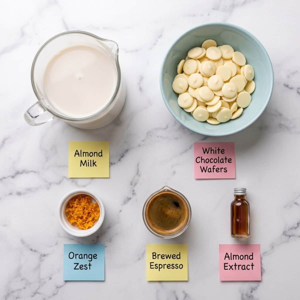 An overhead knolling photograph of almond milk, white chocolate, orange zest, espresso, and almond extract in separate containers on a marble counter with handwritten sticky notes.