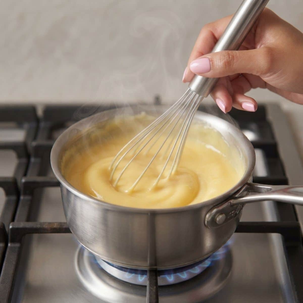 Extreme close-up shot of a woman with baby pink nails whisking thickening custard in a saucepan over a blue gas stove flame.