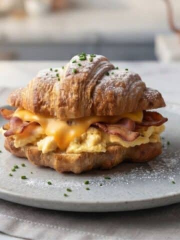 An eye level close-up of Breakfast Croissant Sandwich placed on foggy light blue ceramic plate in a simple American kitchen with natural lighting.