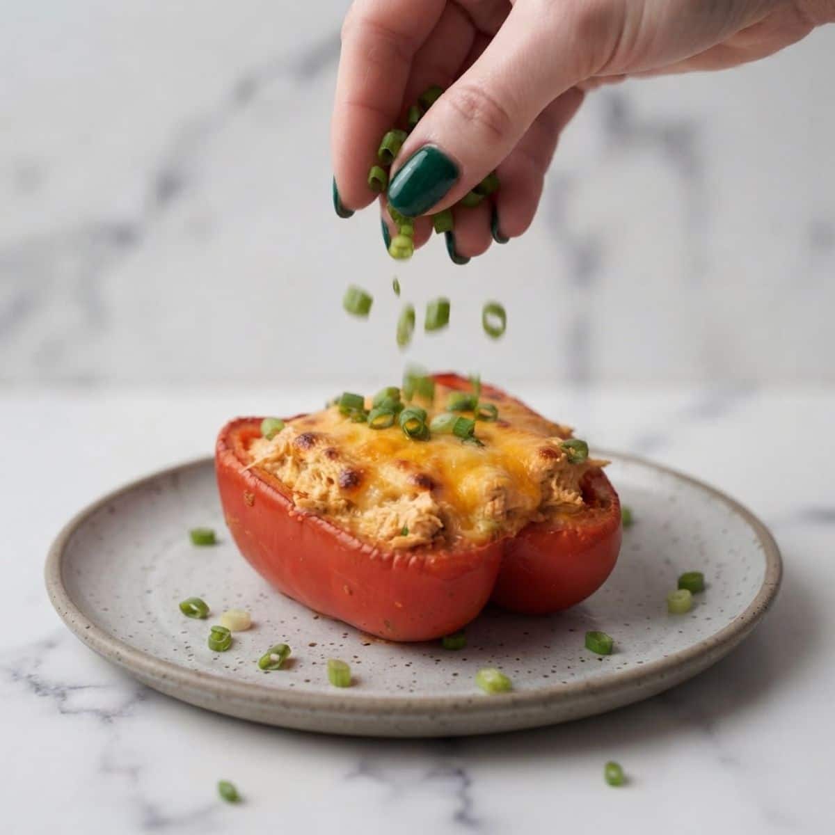 Extreme close-up shot of a woman with dark emerald green nails scattering green onions over a baked Buffalo Chicken Stuffed Pepper on a marble counter.