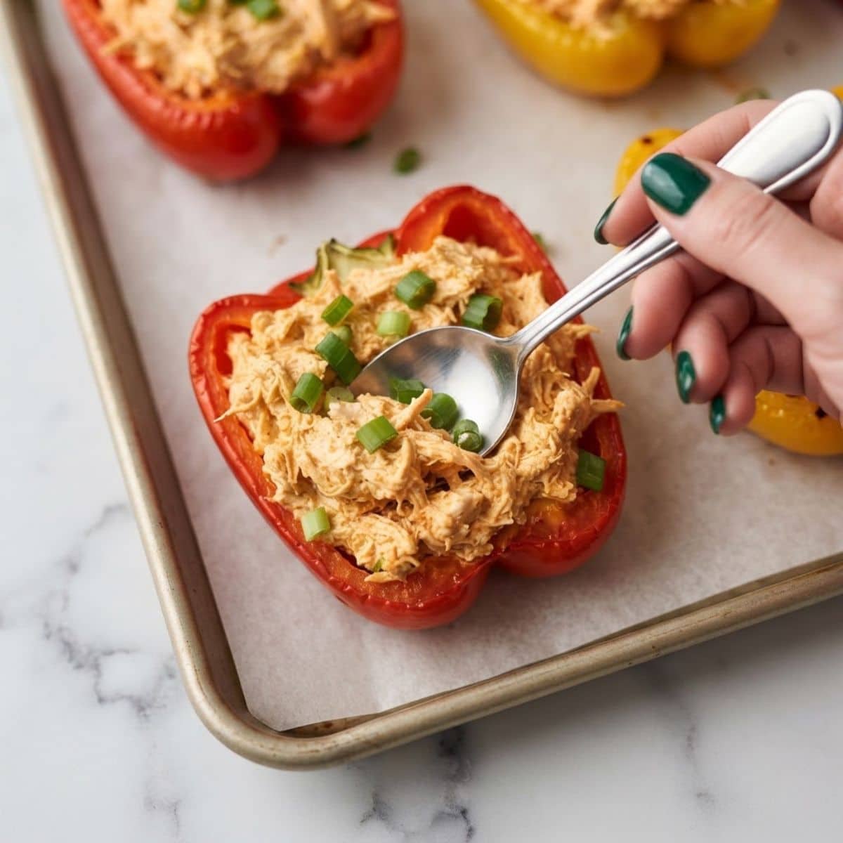 Extreme close-up shot of a woman with dark emerald green nails spooning buffalo chicken filling into a bell pepper half on a baking sheet.